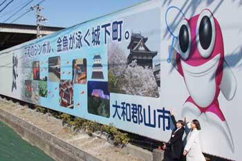 大和郡山市のスローガンと観光名所とピンク色の金魚がモチーフのマスコットキャラクターが描かれた白い看板に手を触れる2人の女性の写真