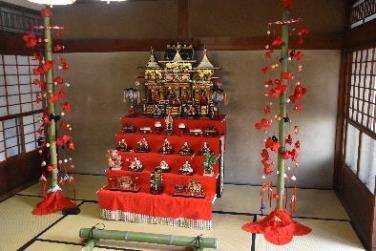 和室にある5段飾りのひな壇とひな壇の両脇にある赤色の飾り物が付いた2本の竹の幹の写真
