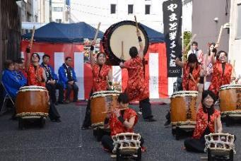 祭りの紅白幕を背に各々大小様々な大きさの太鼓を叩く男女7人の写真