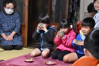 畳の上に敷かれた紫色の布の上で正座の姿勢で両手で茶碗を持つ男女3人の子どもと子どもを見つめる白いマスクをした女性の写真