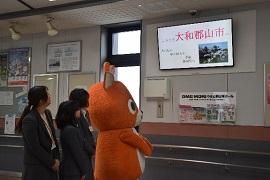 郡山駅内にある「ようこそ大和郡山市へ」の文字と画像が映し出されたモニターを見つめる鹿をモデルにしたオレンジ色のマスコットキャラクターの着ぐるみとスーツ姿の3人の女性の写真