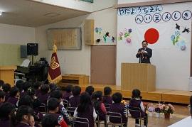 「やたようちえんへいえんしき」と書かれた白く丸い飾り文字と日本国旗が飾られた壁面を背景に壇上でマイクを持つスーツ姿の男性を見つめる着席した園児たちの写真