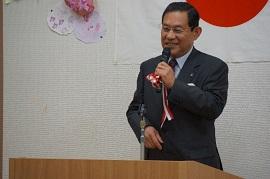 日本国旗が飾られた壁面を背景に演台で紅白の飾りがついた黒いマイクを持つスーツ姿の男性の写真