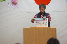 日本国旗が飾られた壁面を背景に演台で紅白の飾りがついた黒いマイクに口を近づけながら両手で赤い屋根の建物のイラストが描かれた紙を持つ白髪の男性の写真