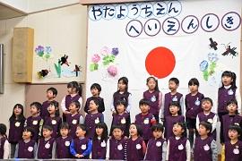 「やたようちえんへいえんしき」と書かれた白く丸い飾り文字と日本国旗が飾られた壁面を背景に壇上に立ち口を開けている紫色のベストを着た園児たちの写真