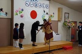 「やたようちえんへいえんしき」と書かれた白く丸い飾り文字と日本国旗が飾られた壁面のある壇上で紫色の大きな旗を握り合うスーツを着た男女2人と男女の園児2人の写真