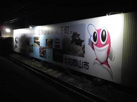照明に照らされた大和郡山市のキャッチコピーや城などの観光名所・ピンク色の大きな目をした魚がモチーフのマスコットキャラクターが描かれた看板の写真