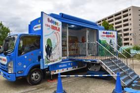 青いトラックの側面が両側に開き扉に鮮やかな絵が描かれ中央にトラックへ上がる階段がある写真