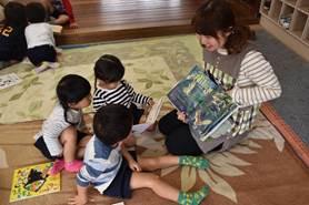 床に座りエプロンの女性が三人の園児に読み聞かせをしている写真