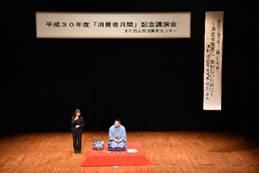 中央に赤い布が敷かれ手前で青紫色の着物をきた笑福亭学光さんが正座し隣で黒いスーツの女性が立っている写真