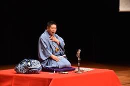 笑福亭学光さんが赤い小舞台の上で正座をして扇子を右手に持って話している写真