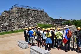 青空のもと、石垣の近くで黄色い帽子の6年生が一か所に集まる写真