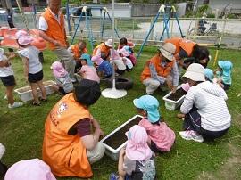 間隔を置いておかれている土が入った植木鉢の周りにしゃがみ込む園児とオレンジ色のベストの大人がいる写真