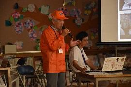オレンジ色のシャツと帽子をかぶった男性がマイクを持って話している写真