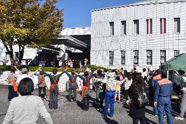 白い建物の前に沢山の人々が集い両手を頭上に広げて立っている写真