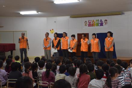 横縞の幼稚園児たちの前で話している揃いのオレンジ色のウェアを着た人々の写真