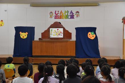 舞台上に置かれてあるキャラクターの飾りがされた藍色の幕と茶色のものを見つめている幼稚園児たちの写真