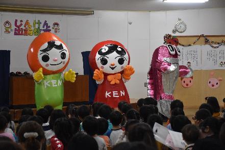 草色と赤色とピンク色の着ぐるみを着た3匹のキャラクターとそれを見つめている幼稚園児たちの写真