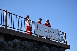 赤い洋服を着た男性2人と赤いスカートを履いた女性が「集え！未来への主張」と書かれた横断幕が掲げられた柵のある高いところで立ち話している写真