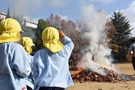 黄色い帽子を被った幼稚園児が燃えているサツマイモの山を眺めている写真
