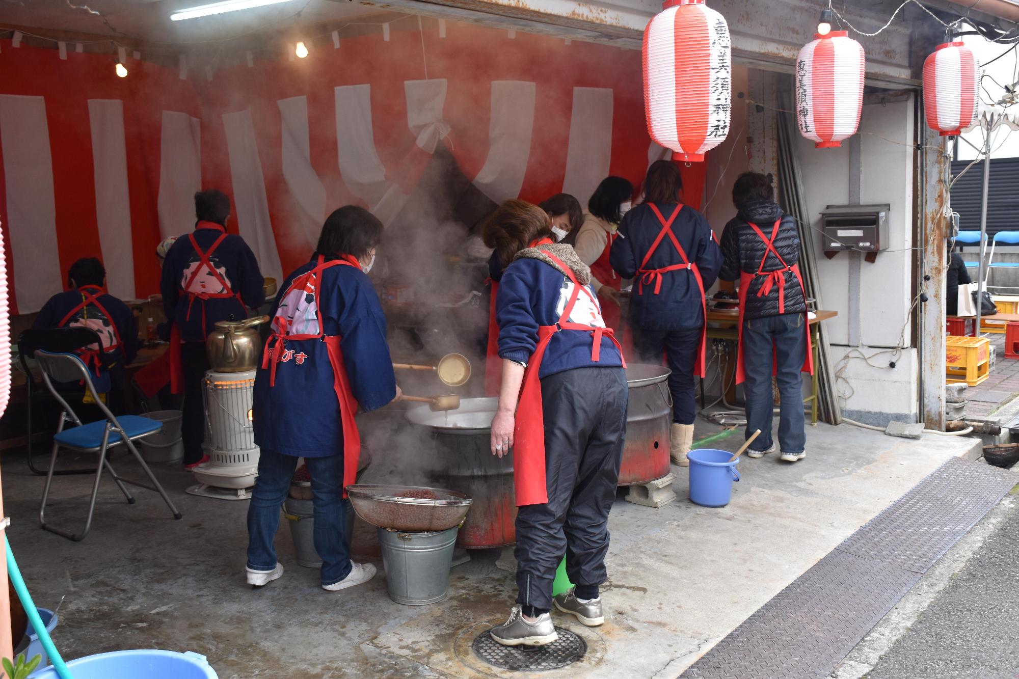 紺色の服に赤エプロン姿の女性たちが大きな鍋で善哉を作っている処の写真