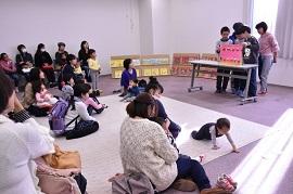 4人組の小学生がピンク色の紙芝居を持って読み聞かせを行っている下で、赤ちゃんがハイハイして歩き回っている写真