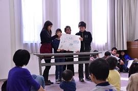 4人組の小学生が手作りの白い紙芝居を持って、親子連れに読み聞かせを行っている様子の写真