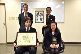 「MOA美術館全国児童作品展」で内閣総理大臣賞を受賞した女生徒と校長が椅子に腰かけて、賞状と記念品をもって撮影された写真