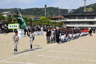 プラカードを持って行進する野球ユニフォーム姿の選手と旗を持つ選手の後に並ぶ選手たちの写真