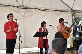 大きなテントの中のステージでギターを弾いている男性と歌っている男女の3人組とそれを聞いている観客の様子の写真