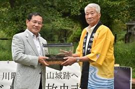 黄色いハッピを着た男性からスズムシの入った箱を手渡しされている市長の様子の写真