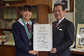 木目壁の市長室にて撮影された表彰された女子学生と市長が二人で表彰状をもった写真