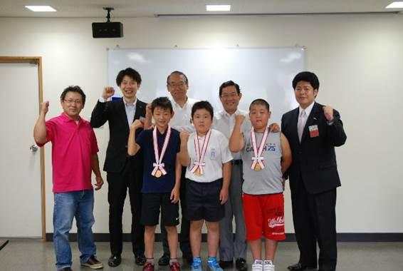 「第23回わんぱく相撲大和郡山場所」で優勝し、全国大会に出場する首からメダルを下げた3人の小学生と5人の大人たちの集合写真