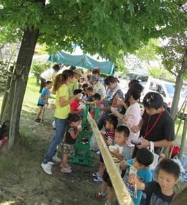 緑がいっぱいの木陰で、竹を挟むようにして並んで立った子供たちや保護者がそうめんを食べている写真