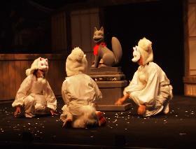 ステージ上の狐の像の周りに集まった狐の衣装をまとった3人の子供たちの写真