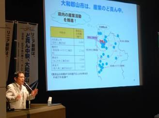 ステージのスクリーン横に置かれたマイクで「リニアをめぐる現状等について」を講演している同盟会会長の上田市長の写真