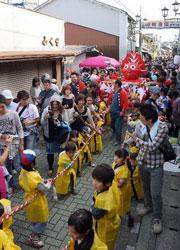黄色いはんてんを着た大勢の子ども達が赤い「ゆるキャラ」が乗った神輿を引いている写真