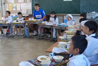 青いユニフォームを着たサッカー選手と給食を食べる小学生たちの写真