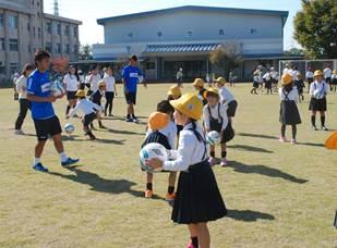サッカー選手にサッカーを教えて貰っている黄色い帽子を被った小学生たちの写真