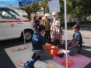 展示された救急車のそばで2人の隊員が、人形を使って救護活動の手順を参加者に教えている処の写真