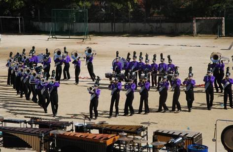 校庭で、紫の衣装を着た大勢の楽団がトロンボーンやトランペットなど色々な楽器を演奏している写真