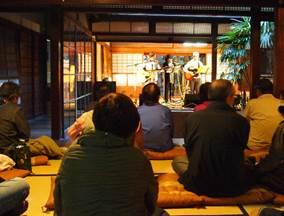 日本家屋で開かれた演奏会を多くのお年寄りが座って聞いている様子の写真
