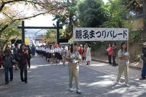 「親子まつりパレード」と書かれた看板を持つ2人の女性と、後ろから白いシャツに黒いズボンを履いた集団が行進している写真