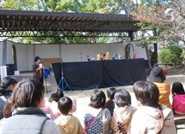 公園の特設会場で人形ショーを開いている人と、それを見に来た子供たちの写真