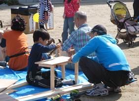青い服を着た男性に教わりながら、金づちで打ち付けながらテーブルを作っている子供の写真