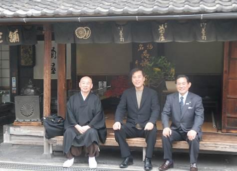 日本建築の軒下で、3人の男性が腰掛け笑顔で記念撮影をしている写真
