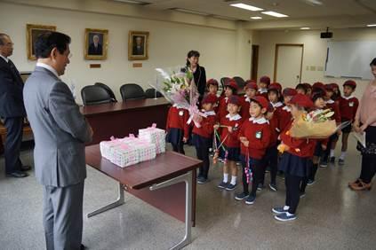 赤い服を着た幼稚園の子らが花束を持って、市長と教育長を訪問した時の写真