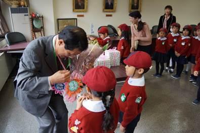 赤い服を着た幼稚園の子らが花束をグレーのスーツを着た男性に手渡す処の写真