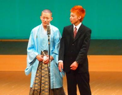 水色を基調とした和服を着た男性とスーツを着たオレンジ色の毛髪の男性が舞台上で横に並びマイクの前に立っている写真