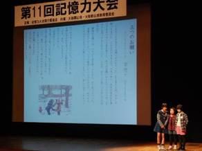 「第11回記憶力大会」と書かれた横断幕の下にある文字とイラストが投影された白いスクリーンのある舞台の脇に立つ3人の女の子の写真